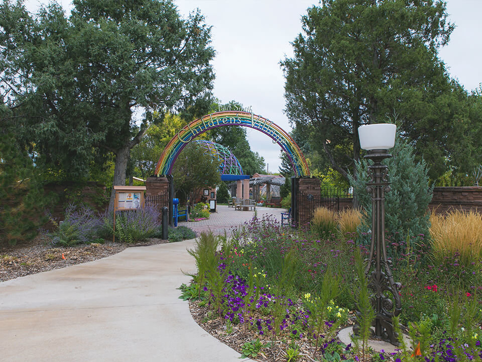 A sidewalk curves past an array of flowers and foliage and bloom, heading beneath a rainbow arch to enter the Paul Smith Children's Village