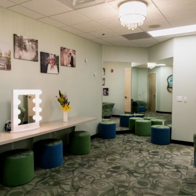A welcoming room with blue and green stools, a lighted mirror, and glowing chandelier.