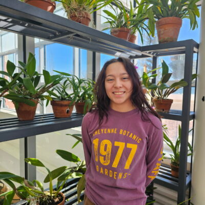 A smiling girl stands in front of several shelves, each holding an assortment of potted plants.
