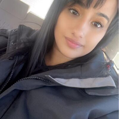 A woman wearing a black jacket sits in her car, smiling.