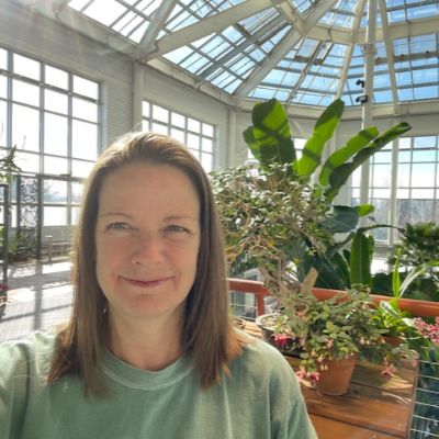 A smiling woman takes a selfie in the Cheyenne Botanic Gardens, showcasing large green leaves behind her.