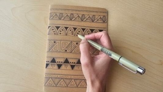 A hand draws geometric patterns on a small brown notebook with a black pen, resting on a light wooden table.