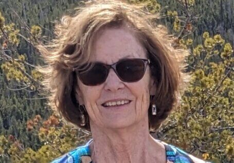 Barbara J. Wolf with short brown hair wearing sunglasses and dangling earrings, smiling outdoors in front of a forested mountain landscape. She is wearing a blue patterned top, and sunlight highlights her face and hair.