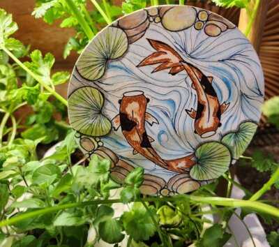 A hand-painted circular artwork featuring two orange, black, and white koi fish swimming through swirling blue water among green lily pads and smooth river stones. The artwork is nestled among bright green herb plants in warm natural light.