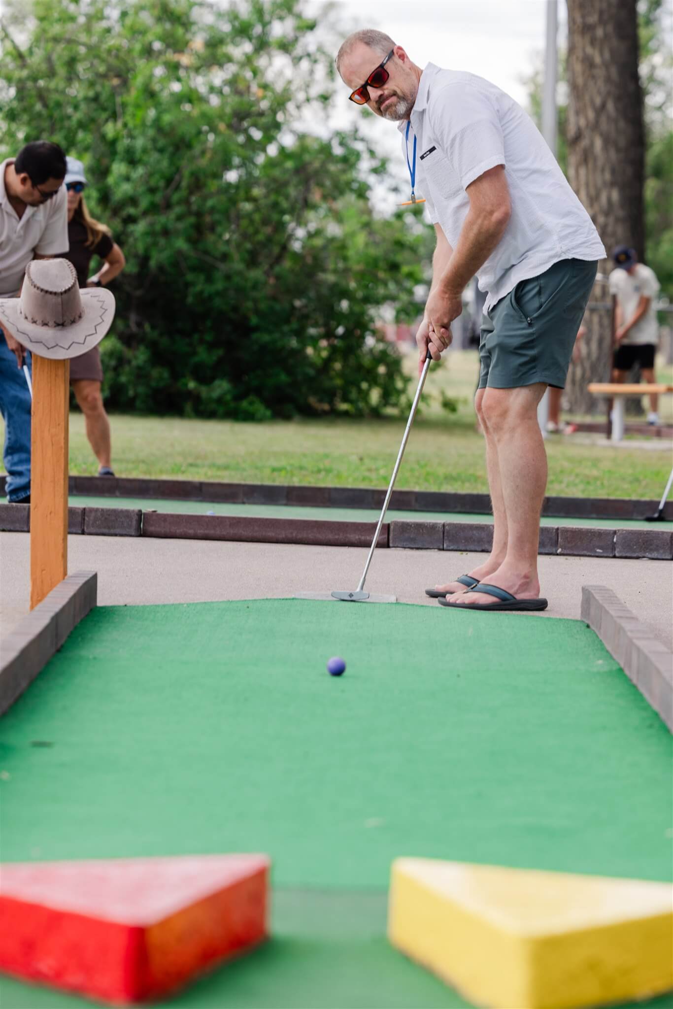 A man wearing sunglasses, a light short-sleeved shirt, shorts, and flip-flops lines up a putt on an outdoor miniature golf course. A purple golf ball sits on the green in front of him, with colorful course obstacles blurred in the foreground.