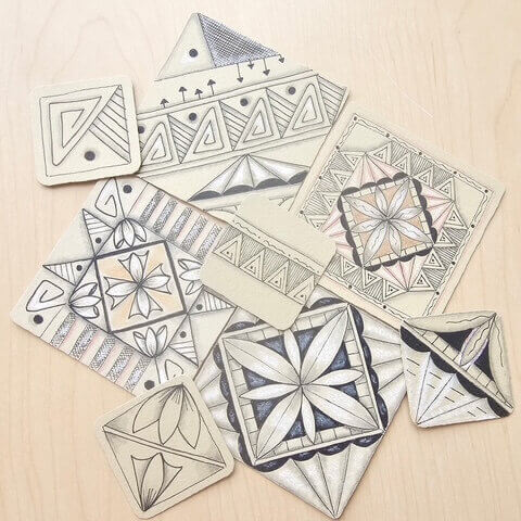 A collection of hand-drawn zentangle-style tiles arranged on a light wood surface. The tiles feature geometric and floral-inspired patterns in black ink with subtle tan, peach, and blue shading. Designs include triangles, petals, stripes, dots, and symmetrical mandala-like shapes on square and diamond-shaped pieces.