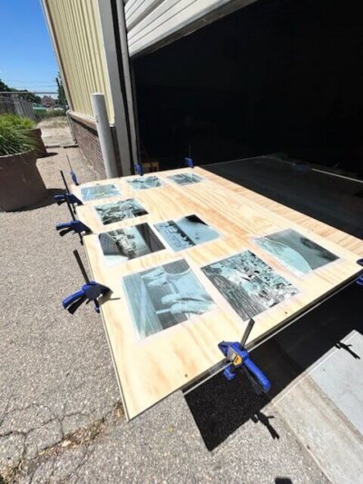 Large plywood panel laid across an open garage entrance with several blue-toned cyanotype prints arranged and secured using blue clamps around the edges. The prints are drying outdoors in bright sunlight on a paved surface beside a tan metal building.